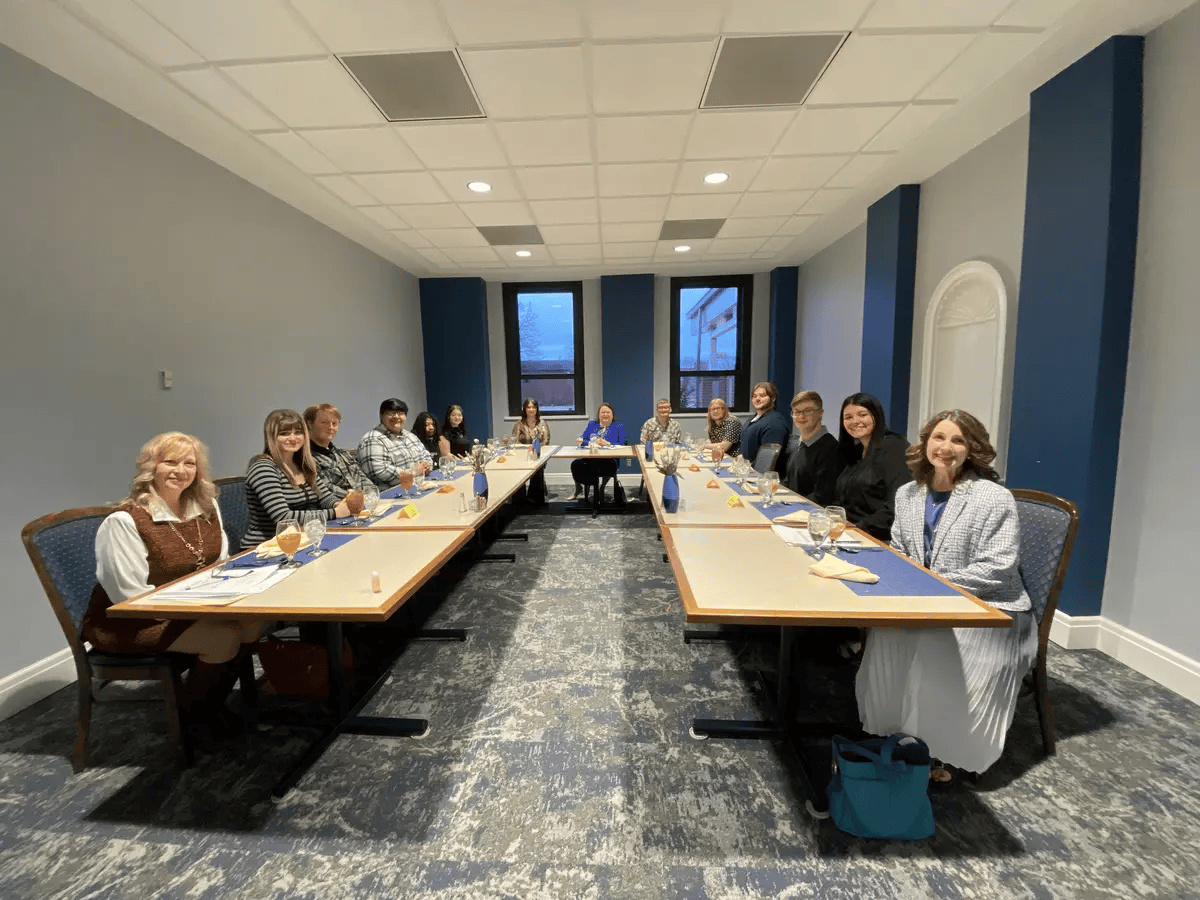 A group of 15 communication club members sit around two long tables in a blue-walled conference room. 