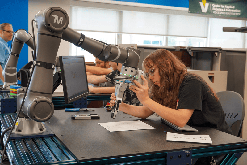 Vincennes University CARA robotics lab: student examines a robotic arm gripper at a lab bench with a laptop.