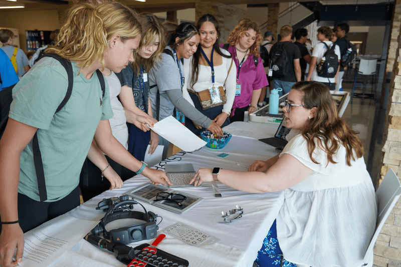 A group of students gathers around a table with tech gadgets, interacting with a woman explaining them.