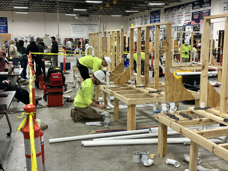 Students practice assembling white PVC pipes and drainage systems beneath wooden mock-up structural frames in a plumbing workshop.