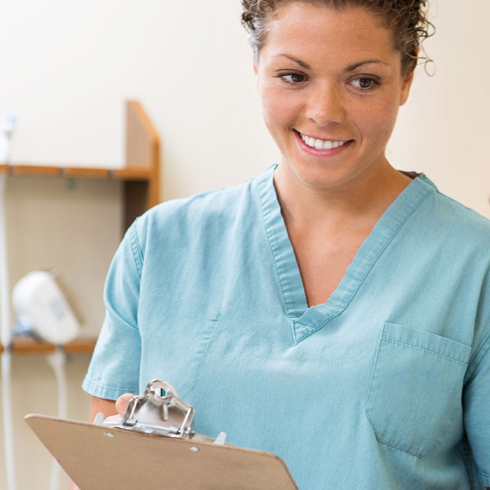 Smiling female healthcare worker in light blue scrubs holds a clipboard in a clinic, conveying warmth, confidence, and professionalism.