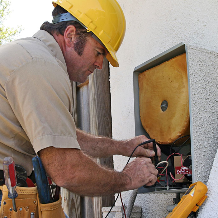 Electrician in a yellow hard hat carefully repairs an outdoor electrical panel, handling wires and tools beside a house wall.