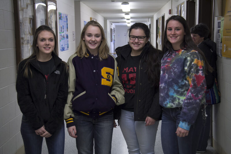 Four young women stand in a school hallway, smiling at the camera. They wear casual outfits, creating a relaxed and friendly atmosphere.
