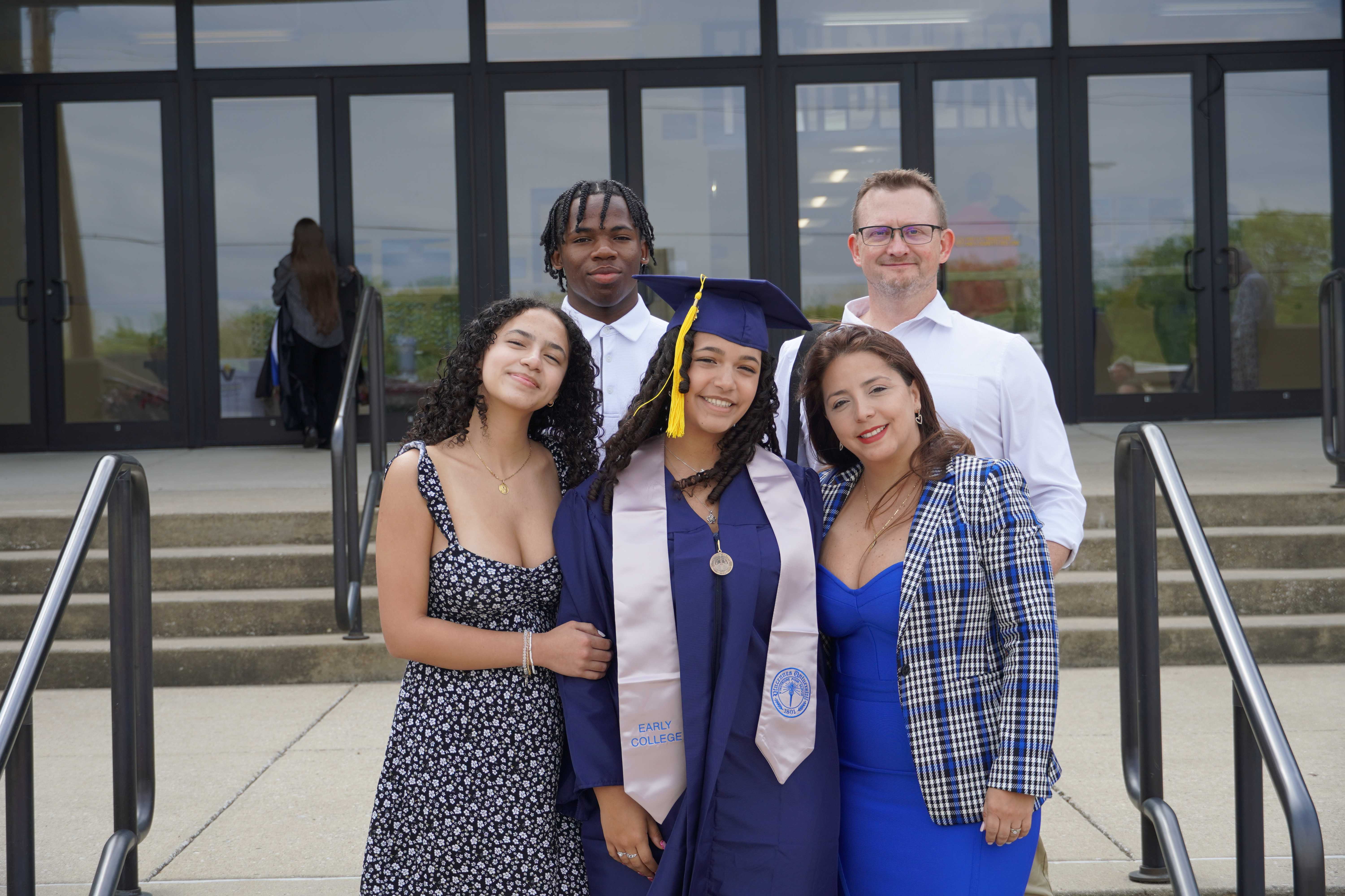 A smiling family of five poses together outside a graduation venue to celebrate a young woman in a purple cap and gown.