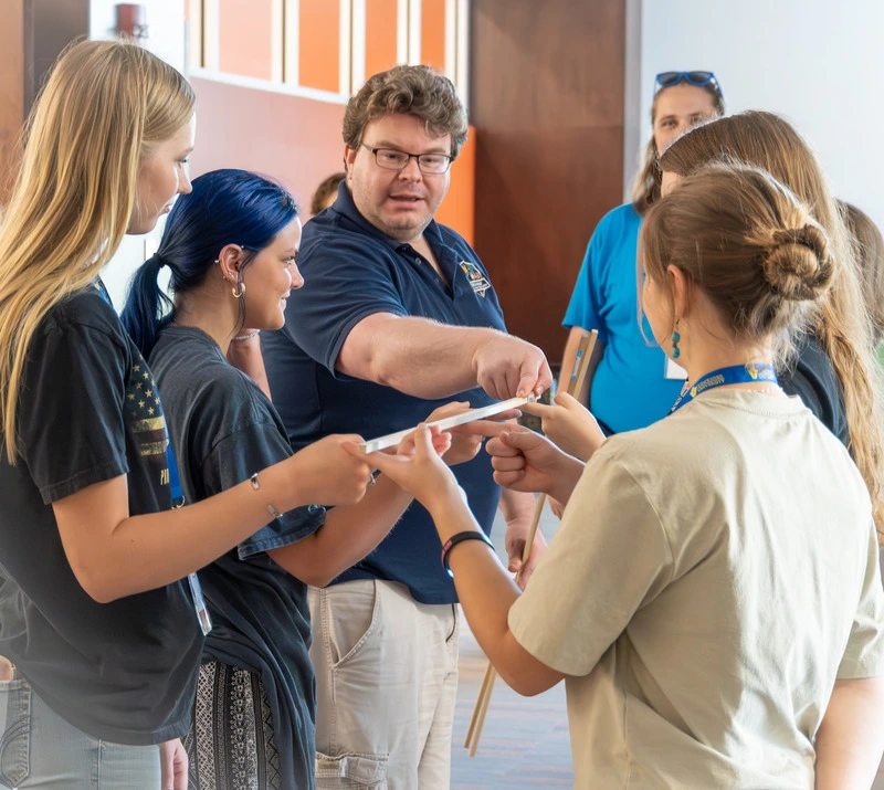 A group of students and a teacher are gathered in a classroom, smiling and interacting.