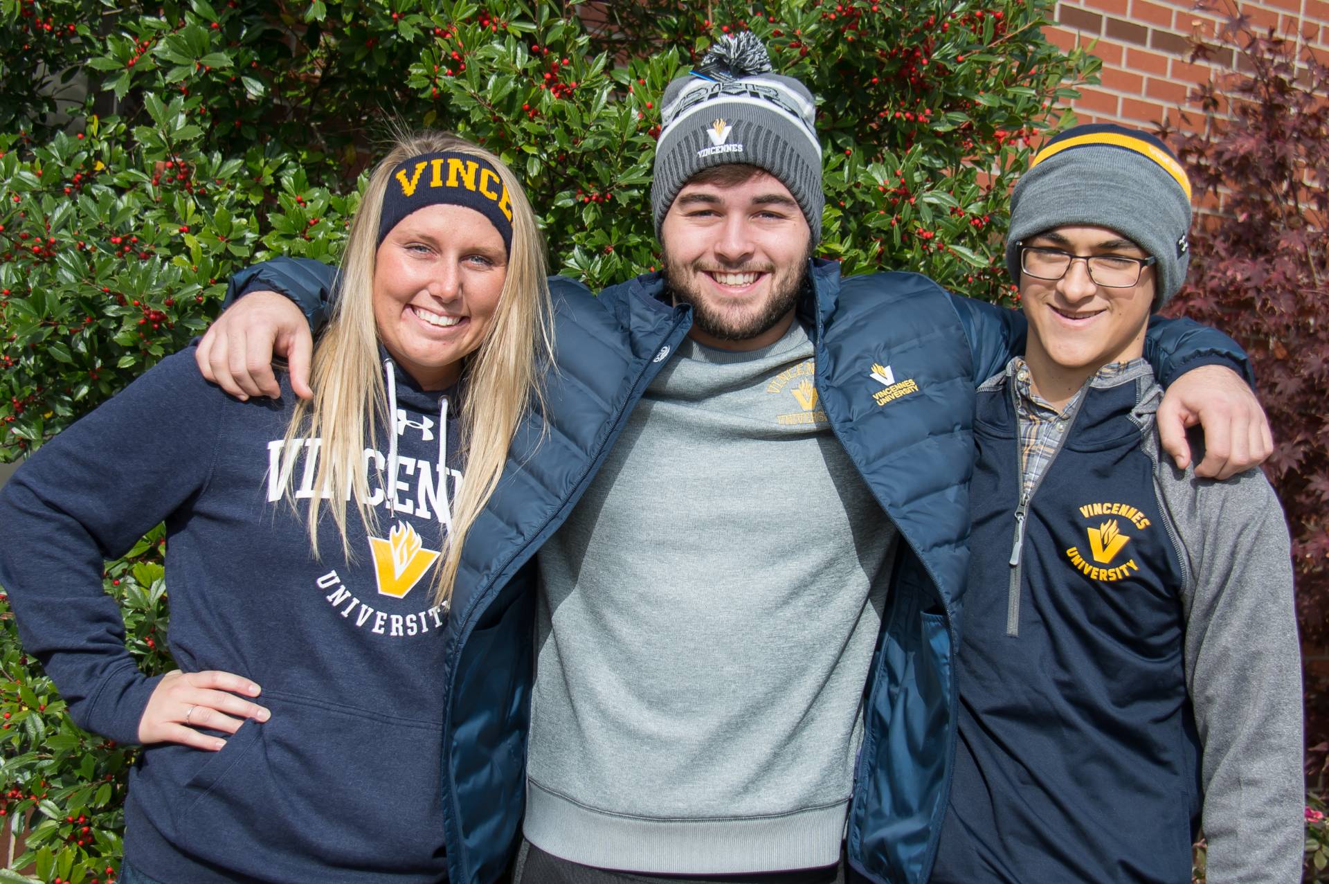 Vincennes University students with arms around each other outdoors, wearing hoodies and beanies, greenery and brick wall in background.