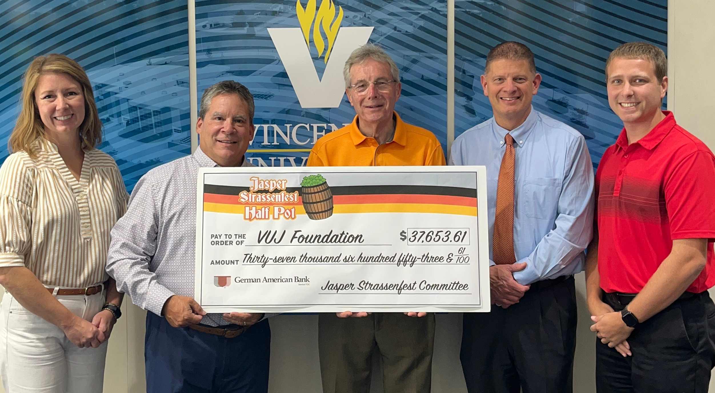 Five people pose while holding a large $37,653.61 presentation check from Jasper Strassenfest to the VUJ Foundation in front of a VU logo.