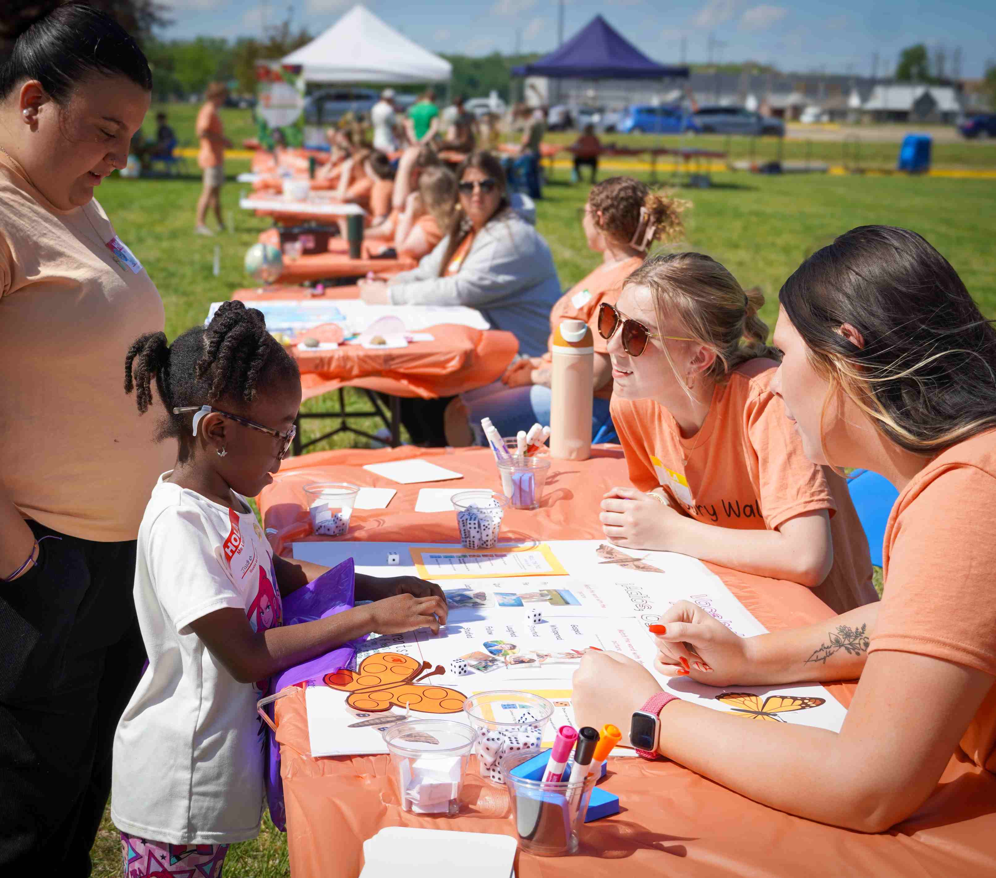 A young girl interacts with VU students in orange shirts at an outdoor event activity table lined with learning games.