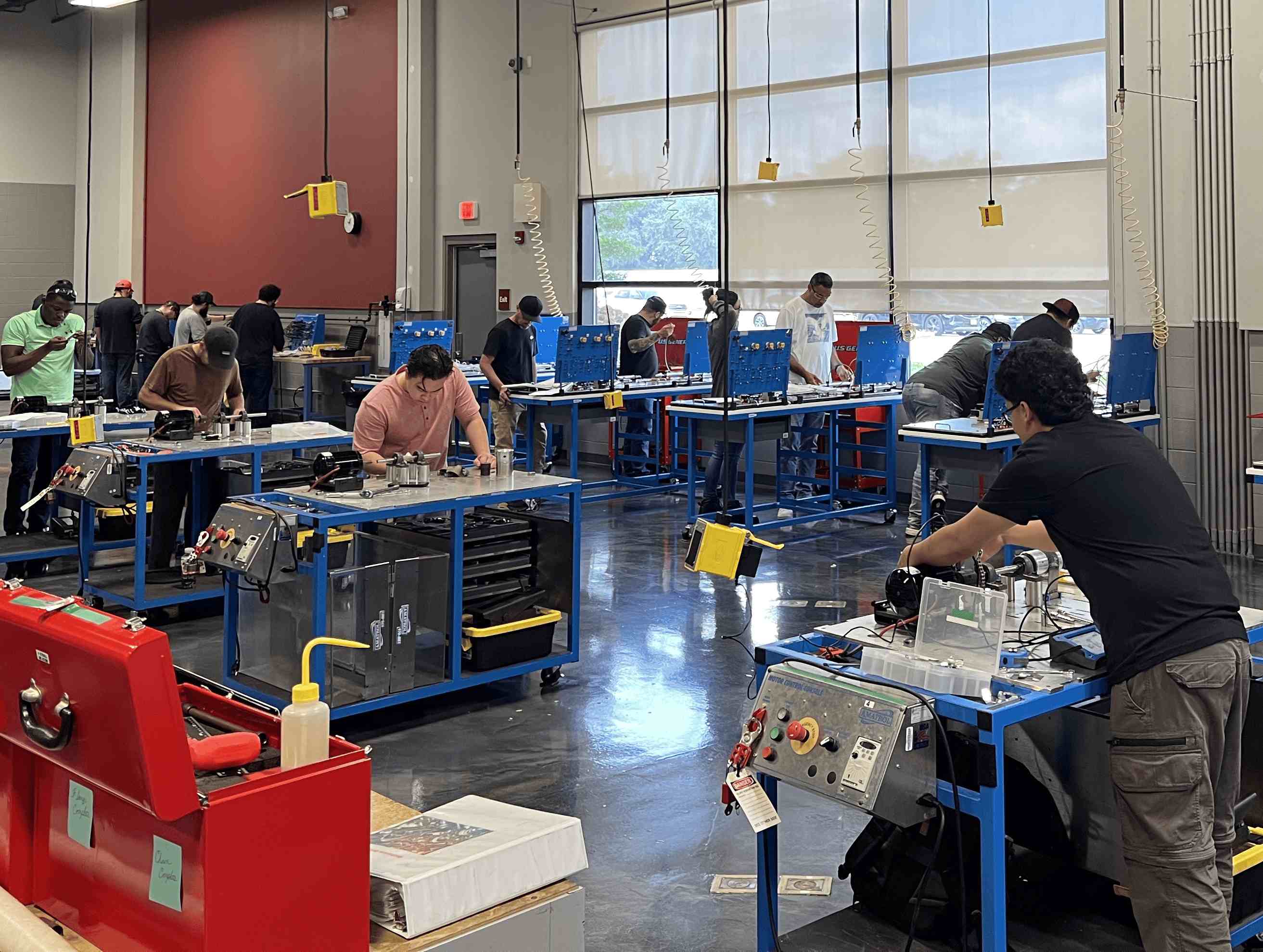 A spacious technical training classroom filled with students working at blue industrial workbenches with mechanical and electrical components.