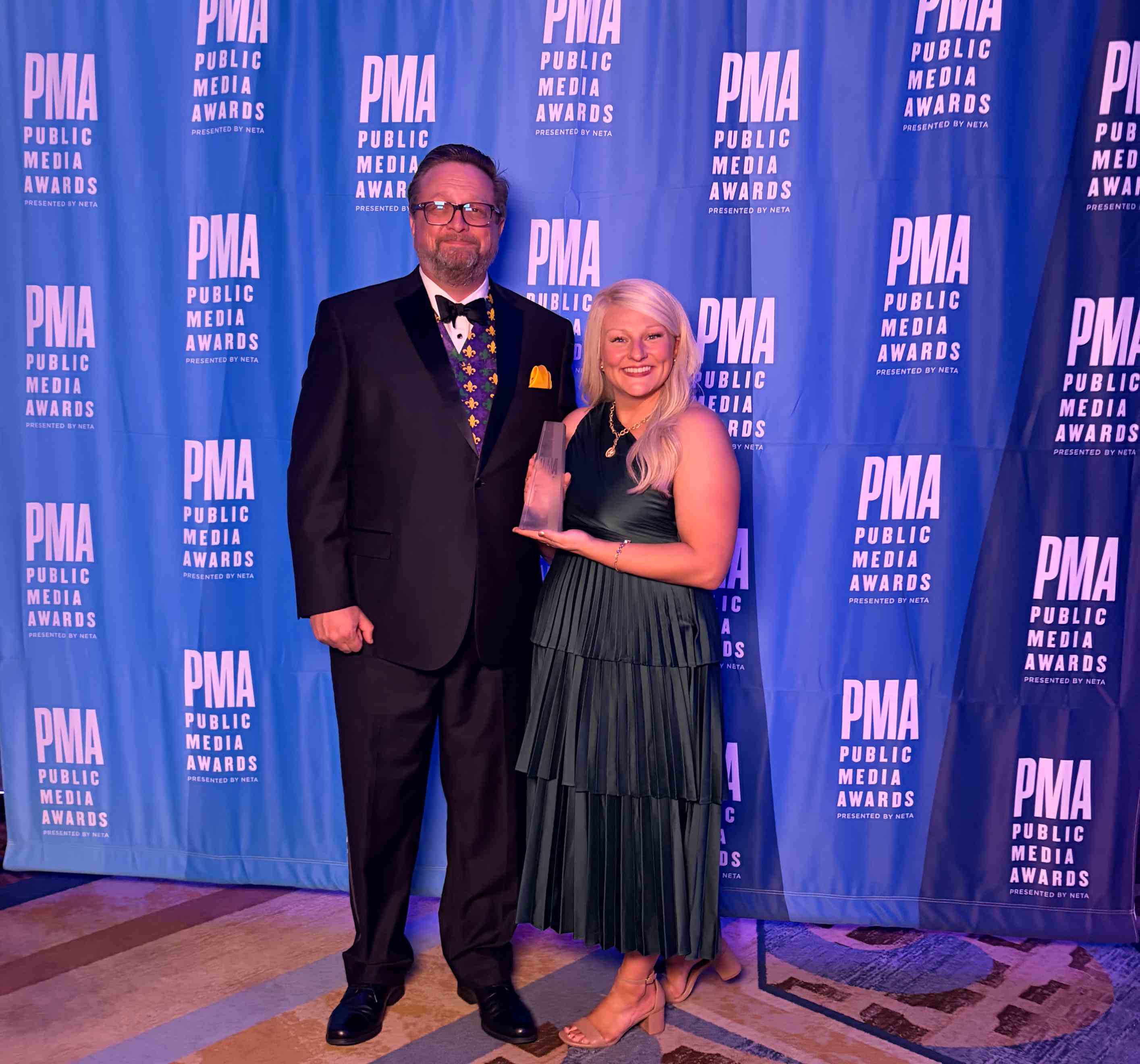 Allyson Bowers wearing a formal dress and smiling holds a Public Media Award trophy, standing beside a man in a tuxedo before a blue PMA backdrop.