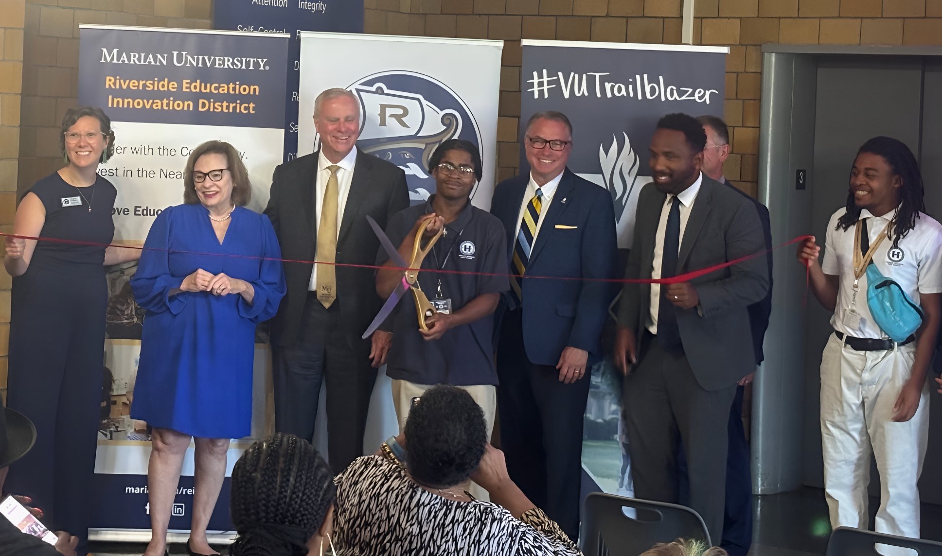 As student cuts a red ribbon with large ceremonial scissors, surrounded by smiling university and high school leaders standing in front of event banners