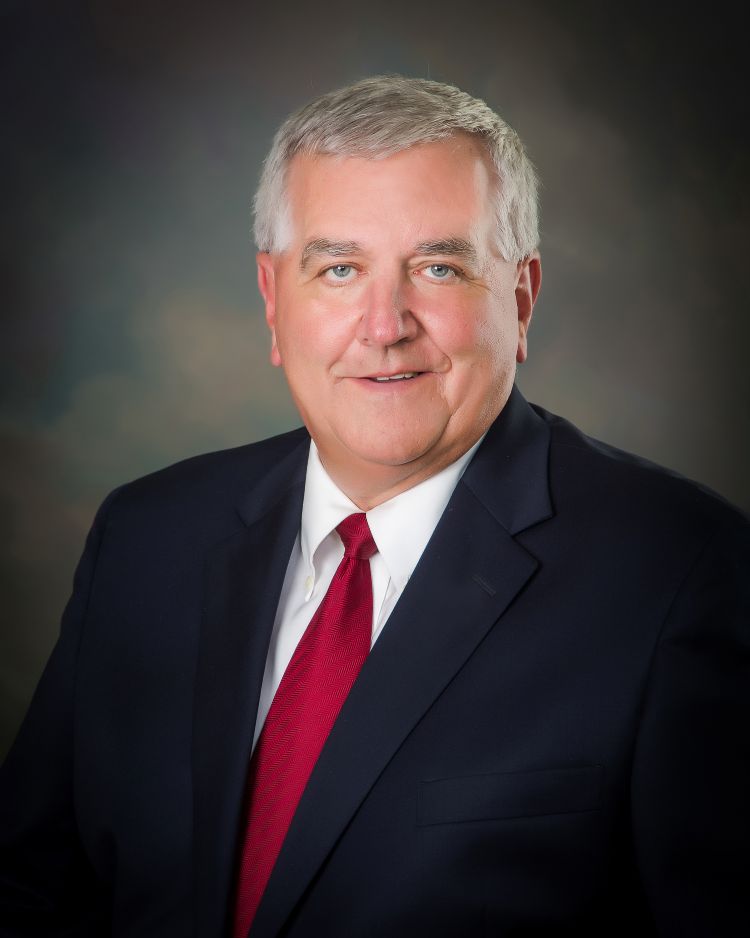 Headshot of a smiling, Kirk Bouchie wearing a dark blue suit jacket, a white collared shirt, and a red tie.