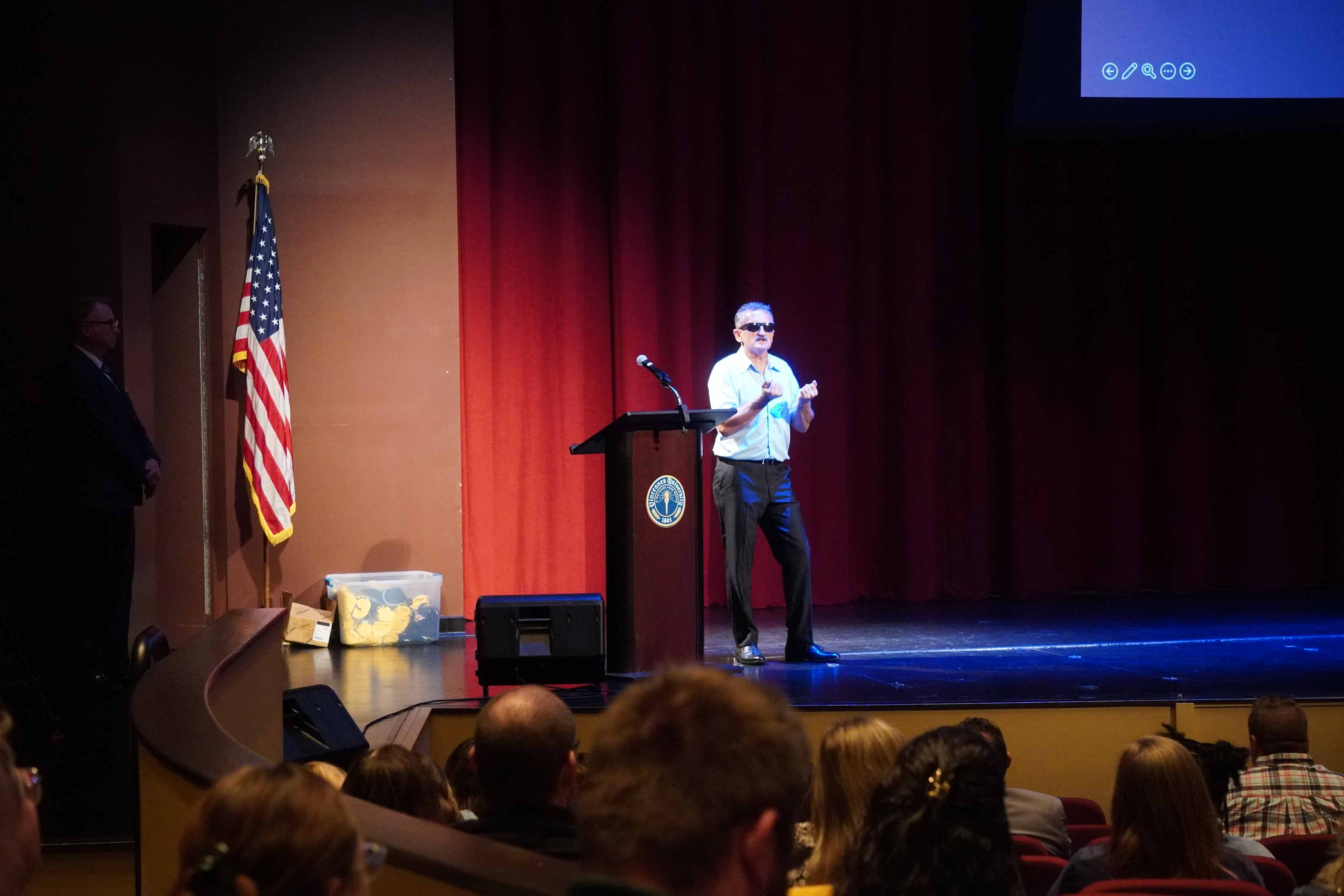 Lonnie Bedwell, a man wearing sunglasses, speaks on a stage beside a podium and an American flag, addressing an audience at the Red Skelton Performing Arts Center..