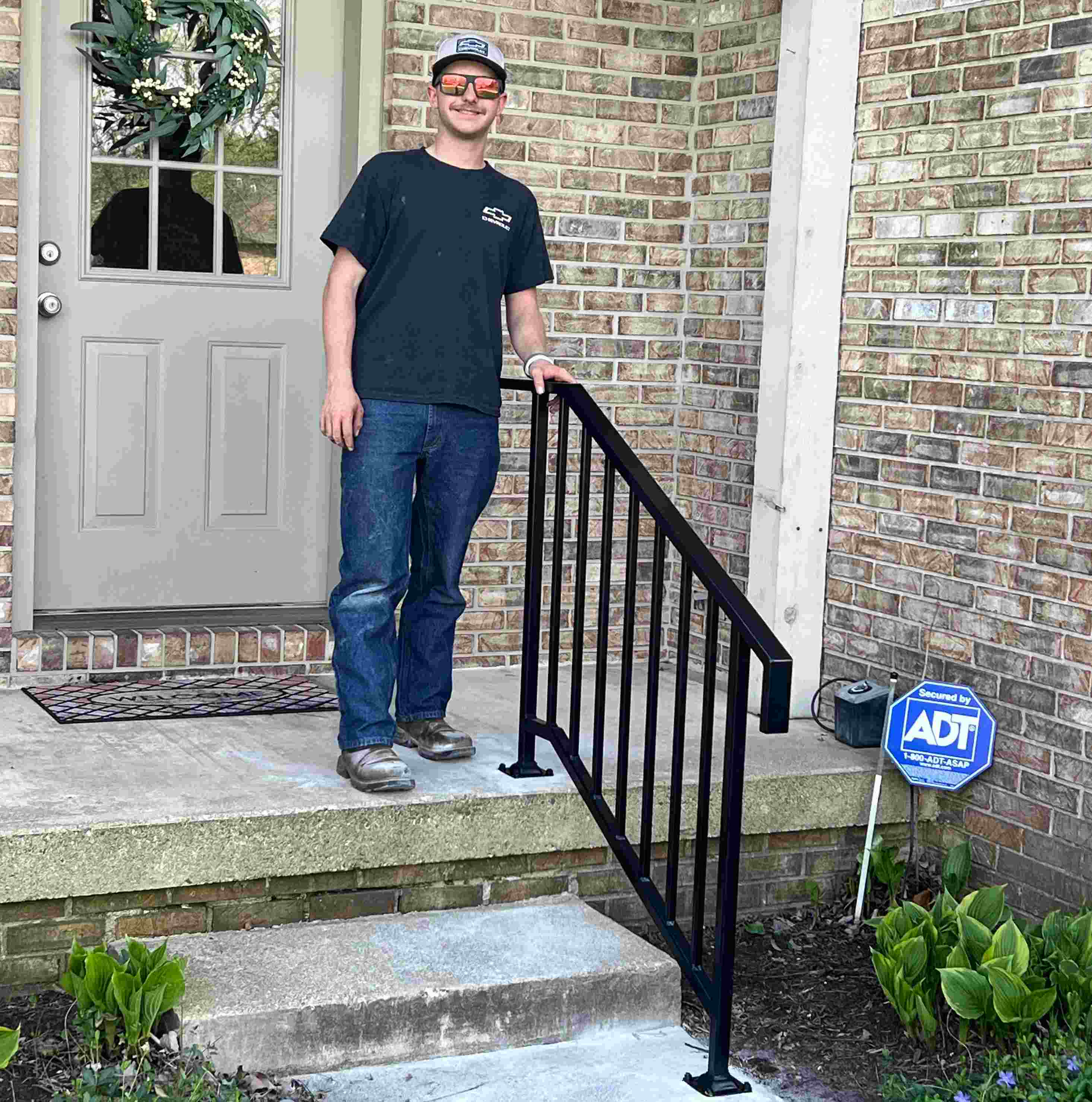 Owen Hart in a cap, sunglasses, black t-shirt, and jeans stands on concrete porch steps, resting his hand on a black metal railing.