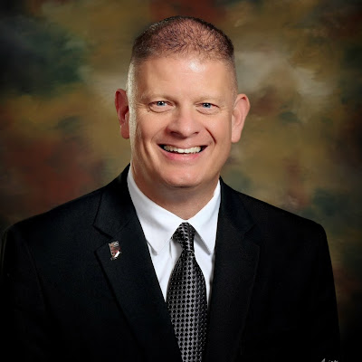 Headshot of Travis Madison, a smiling man with short hair wearing a black suit jacket, white dress shirt, and patterned tie against a studio backdrop.