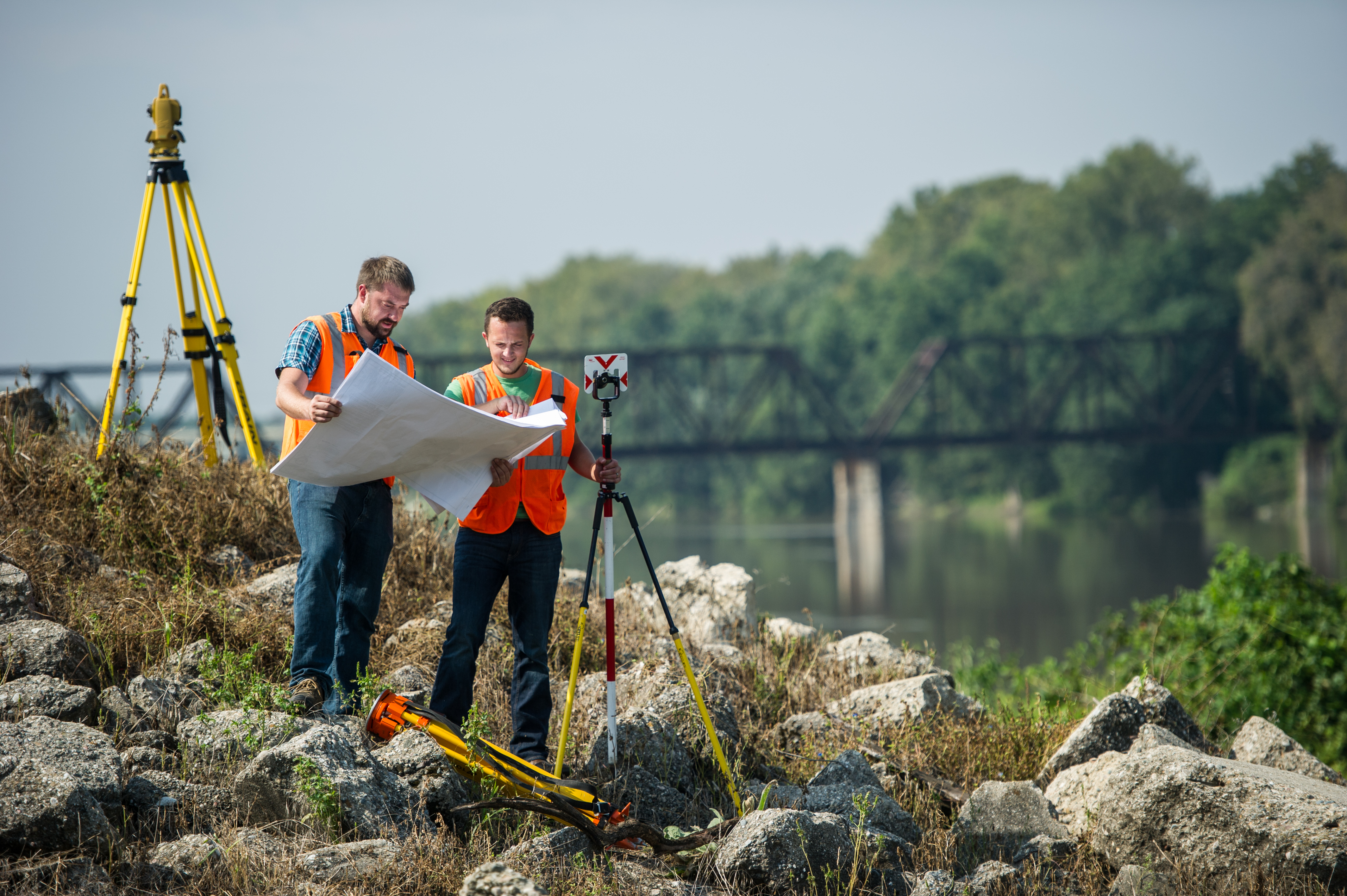 Two males in safety vests inspect a site map outdoors near surveying equipment and a river with a bridge.