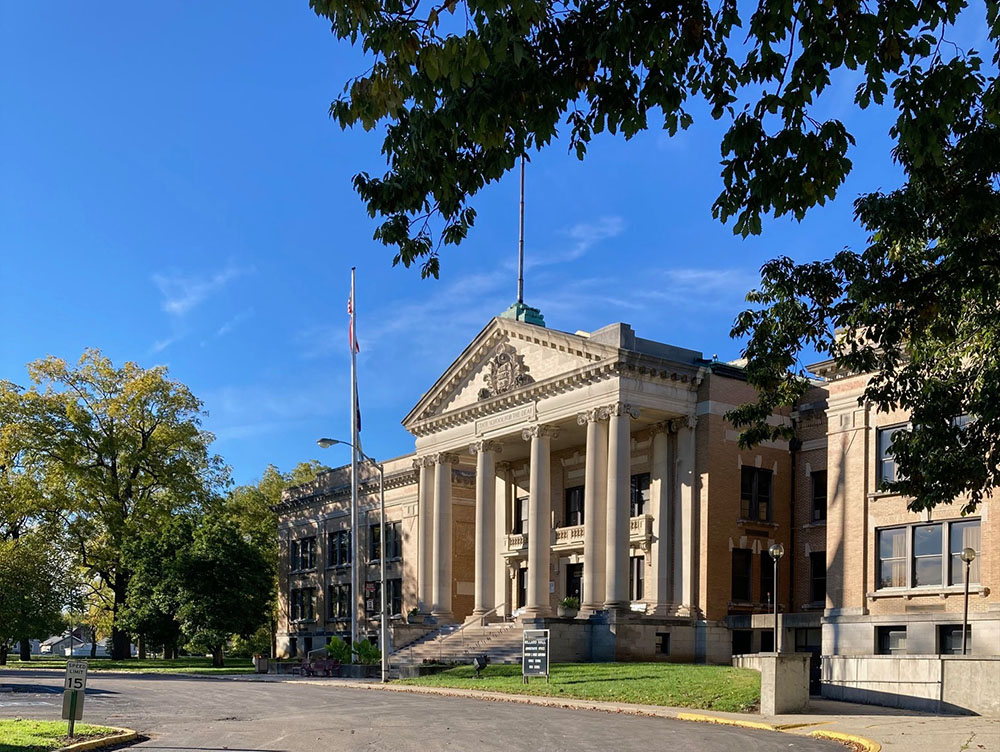 Exterior view of the Indiana School for the Deaf, a Beaux-Arts-style building. The building features a prominent portico with classical columns and a triangular pediment. An American flag waves from a pole to the left of the main entrance. Green grass and trees surround the building, and a paved road leads up to it. The sky is a clear blue with wispy clouds. Tree branches frame the top of the image.